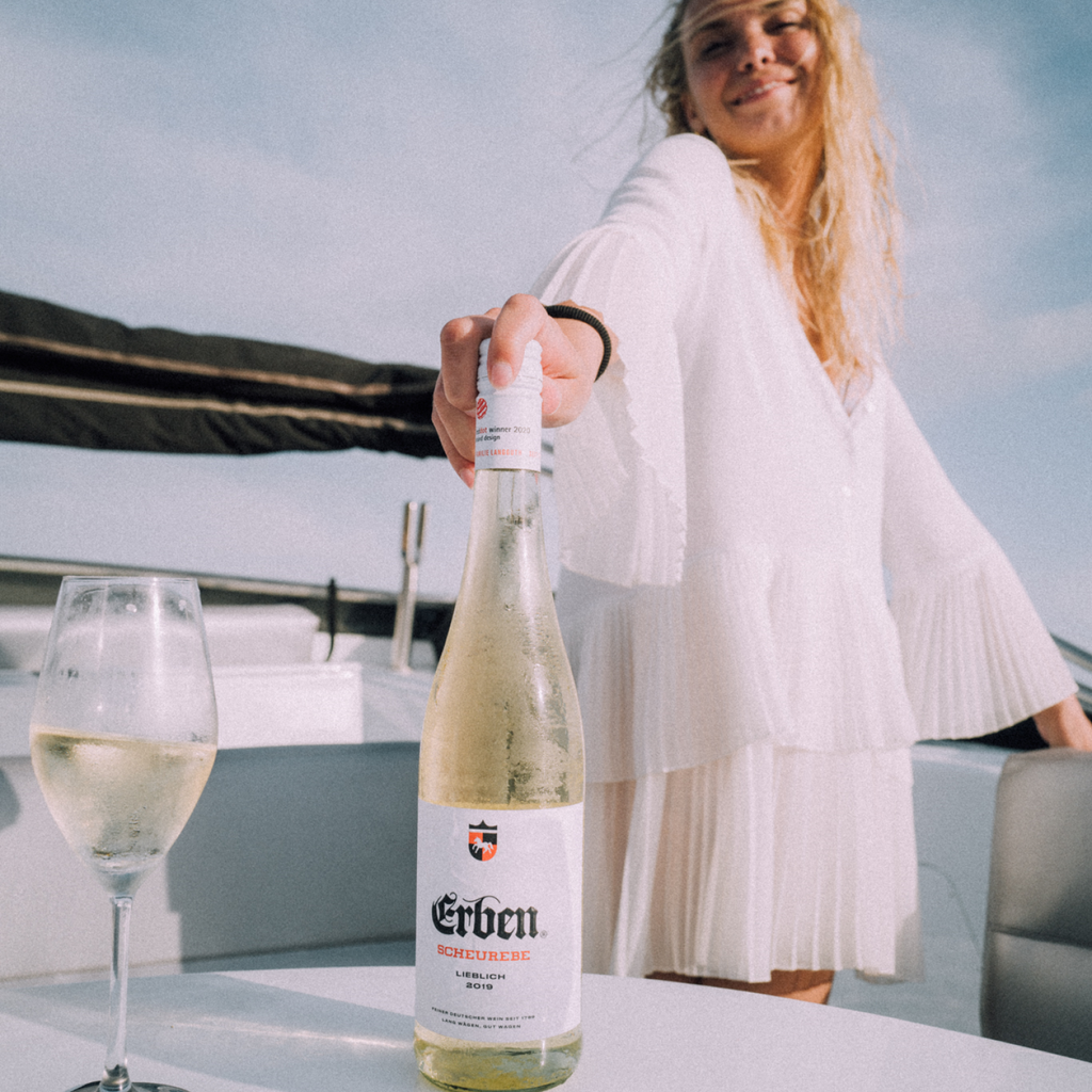 ERBEN Scheurebe Lieblich 0,75l - Weißwein - stehend auf einem Tisch, im Hintergrund eine Frau in einem weißen Kleid 