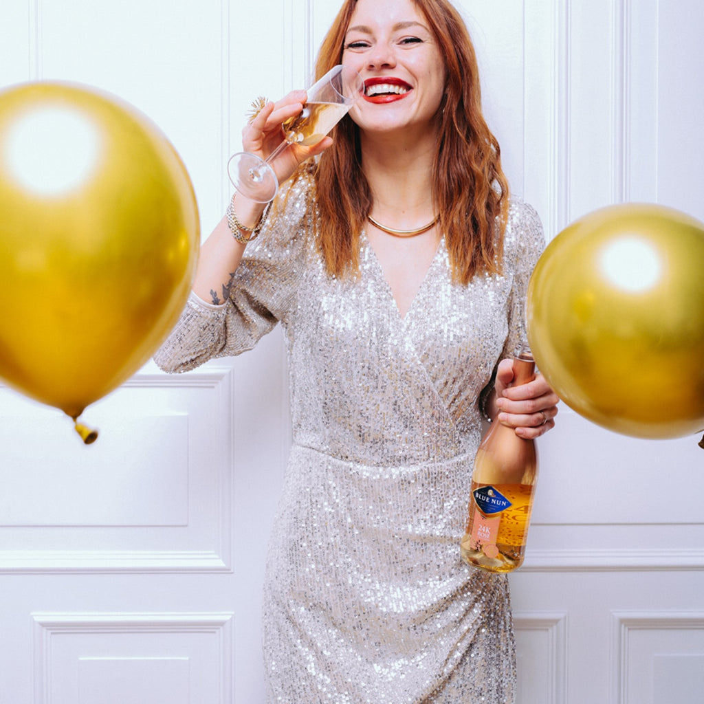 Junge Frau hält ein Glas mit 24K Rosé Gold Sparkling in der einen Hand, in der anderen eine Flasche Blue Nun 24K Rosé Gold